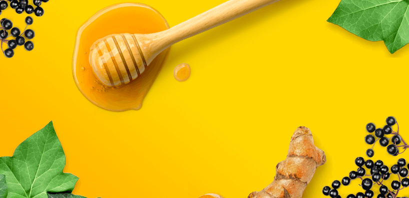 Du miel, des feuilles de lierre, de baies de sureau et une racine de curcuma sur une surface jaune.